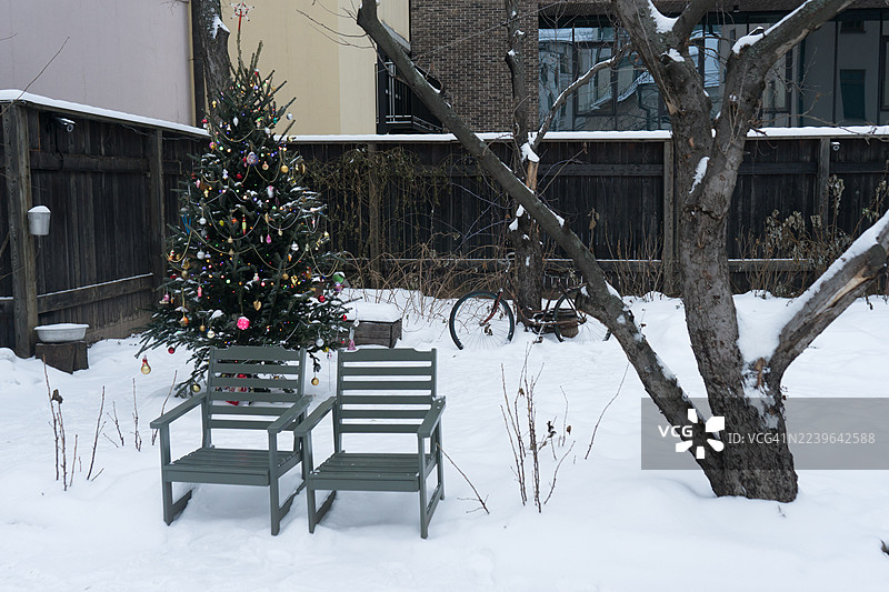 覆盖着雪的后院里有两把空椅子，旁边还有一棵装饰过的圣诞树和一辆旧自行车。图片素材