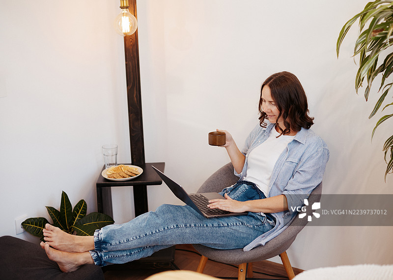 放松的成年女性在家中用笔记本电脑工作，手中拿着咖啡，周围是植物装饰。图片素材