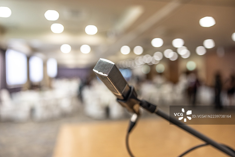 前景中的麦克风支架清晰对焦，模糊的背景中是会议室，有桌椅和舞台，显示已为活动或演示做好准备。图片素材