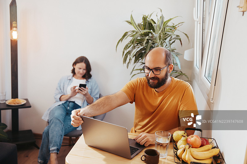 成年情侣在厨房餐桌旁用笔记本电脑在家办公，旁边有新鲜水果和植物。图片素材