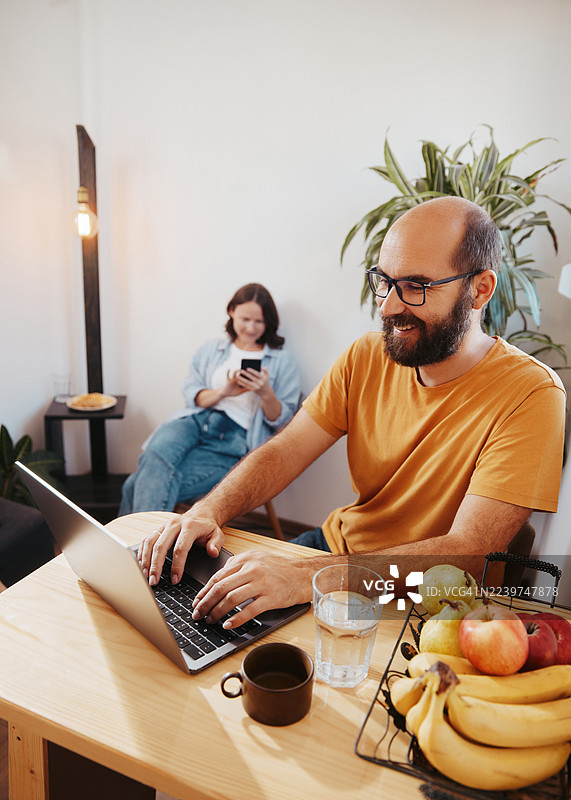 一对夫妇在厨房餐桌旁用笔记本电脑在家办公，旁边放着新鲜水果和植物，他们的家很小，但很温馨。图片素材