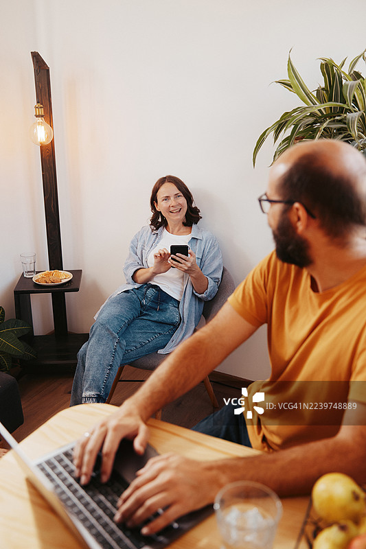 一对成年夫妇在厨房的桌子旁，用笔记本电脑在家办公，旁边有新鲜水果和植物。图片素材