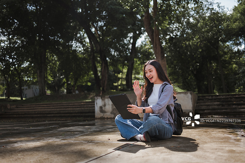 年轻的亚洲学生坐在地上，在公园里通过平板电脑进行在线视频通话，远程学习，并挥手致意。图片素材