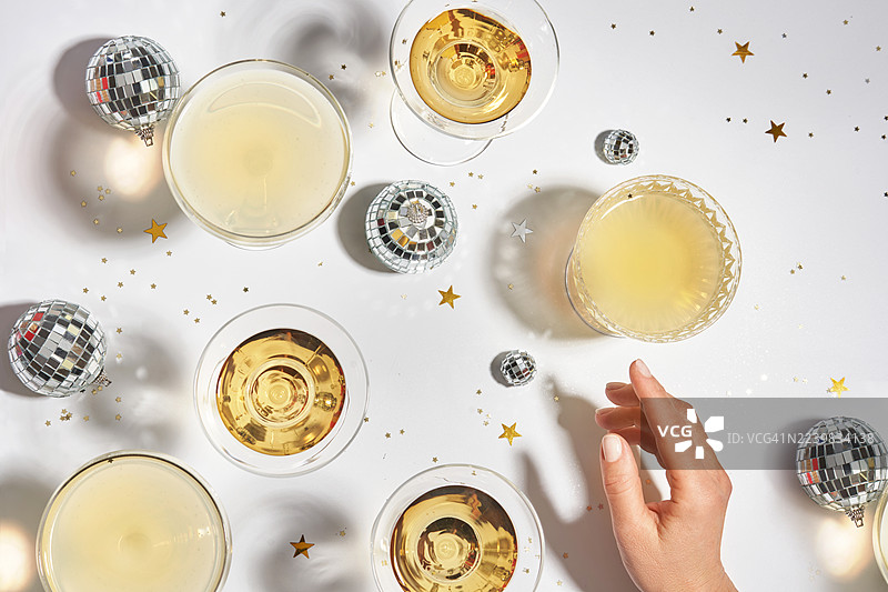节日新年派对上，一只带有自然美甲的女性手正在伸向一杯酒精饮品。圣诞餐桌布置，摆放着马提尼、玛格丽塔鸡尾酒杯、香槟、葡萄酒，背景为白色，上面装饰着银色迪斯科球。图片素材