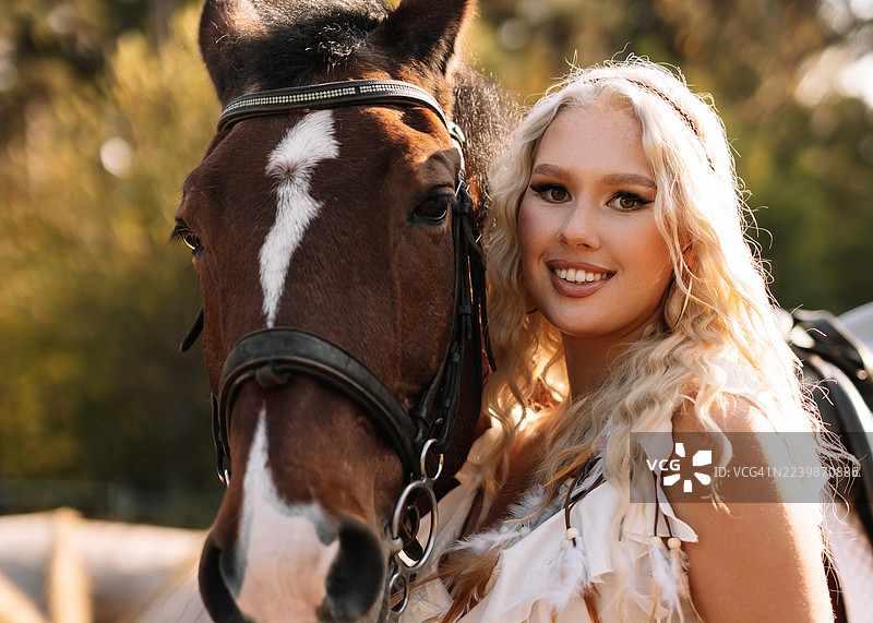 漂亮的金发女郎戴着牛仔帽，在乡村的牧场或马厩里与马站在一起图片素材