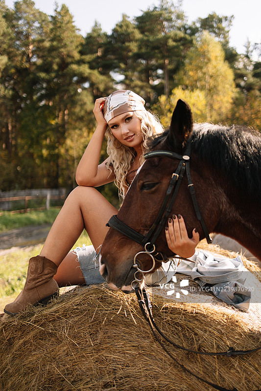 一位美丽的金发女郎戴着牛仔帽，站在乡间牧场或马厩的马旁。图片素材