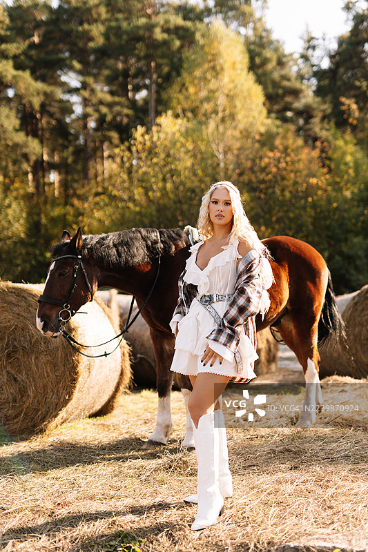 乡村牧场或马厩里，戴着牛仔帽的漂亮金发女郎与马站在一起图片素材