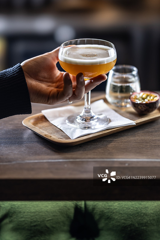 一位女士端着一杯泡沫顶的优雅鸡尾酒——“马提尼”，配有半个百香果和一小杯气泡酒，盛放在木质托盘上。图片素材