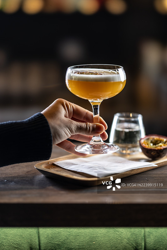 一位女士在木质托盘上拿着一杯优雅的马提尼鸡尾酒，上面有泡沫，配有半个百香果和一杯气泡酒。图片素材