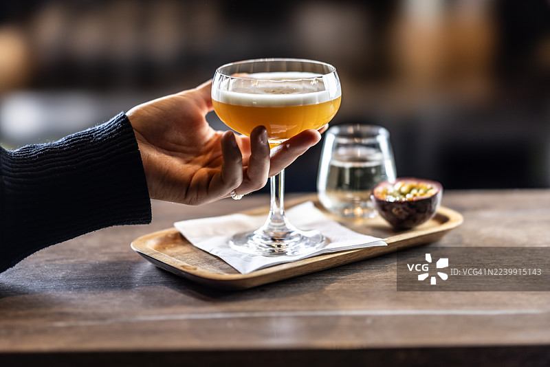 一位女士拿着一杯带有泡沫顶的优雅鸡尾酒——“风情百事达”，配有半个百香果和一小杯起泡酒，盛放在木质托盘上。图片素材