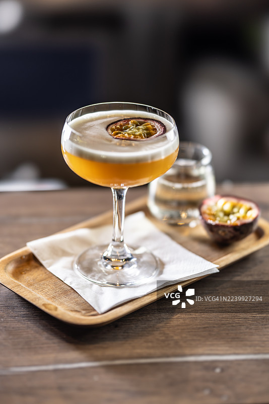 酒馆或夜店吧台上，一杯泡沫丰富的性感尤物马提尼鸡尾酒，用百香果装饰，旁边放着一小杯普罗塞克或香槟，盛放在木质托盘上。图片素材