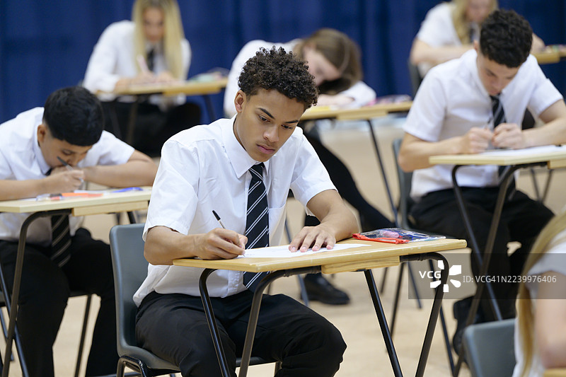 高中男生在考试时坐在书桌前，在小册子上写字，表情专注图片素材