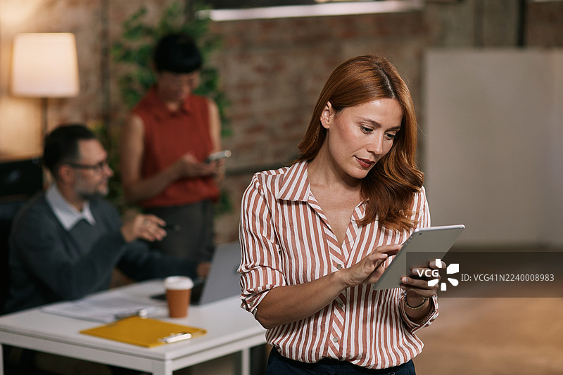 年轻貌美的女商人，手持平板电脑，背景是现代办公室的同事们图片素材