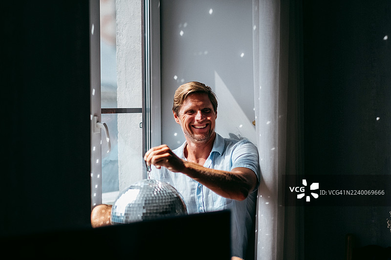 男人坐在窗台上，对着迪斯科球和窗户微笑。派对氛围、室内灯光效果和休闲概念。图片素材