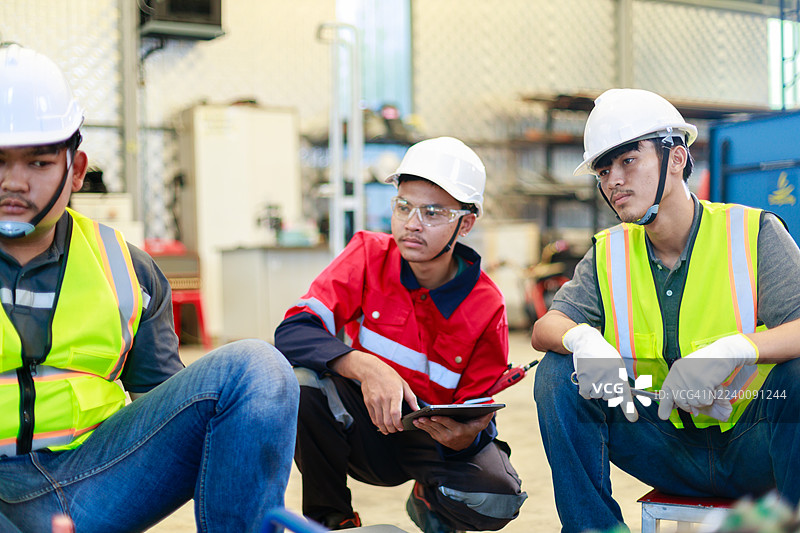 三名身着安全防护装备的工程师在工业车间内，使用液压对接焊机讨论HDPE管道连接，重点展示团队合作、规划和管道安装流程。图片素材