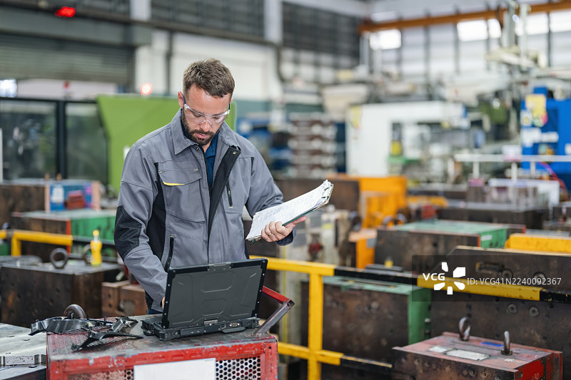 一名男性技术人员在工厂工作，他手持文件并使用笔记本电脑。专业人士在工厂工作。图片素材