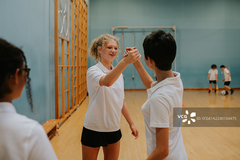两个女孩在上体育课时击掌。一个女孩是一个高加索人，另一个是亚洲人。她们都穿着运动服。她们在学校体育馆里。有其他学生在背景中，但她们是模糊的。她们都在微笑，看起来很高兴。这个场景发生在白天，室内光线充足。图片素材