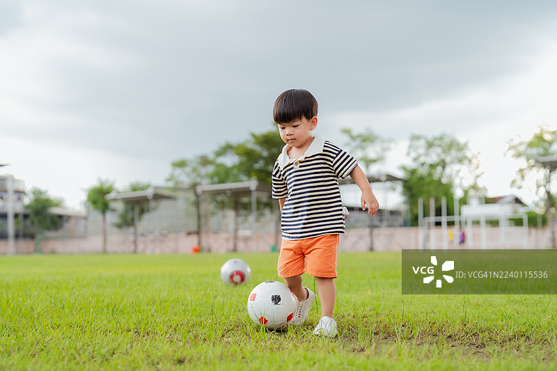 快乐的亚洲小男孩在绿茵场上踢足球，在享受户外运动和健康生活方式的同时，发展平衡力、协调性和体能，脸上洋溢着喜悦的笑容。图片素材