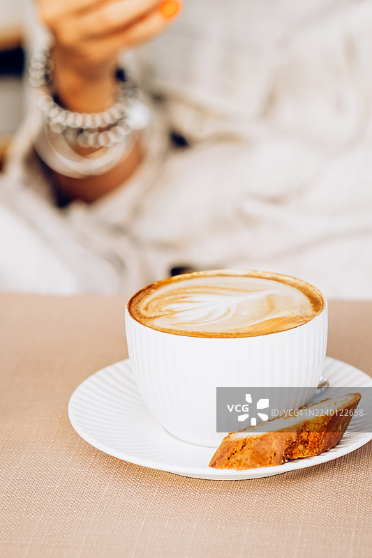 卡布奇诺配比斯考蒂饼，背景是模糊的女士。咖啡文化、放松时刻和舒适氛围的概念。图片素材