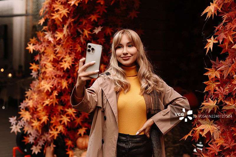 一位年轻的女网红在秋天街道上，背景是橙色的落叶，她正看着手机自拍。一位Z世代女孩穿着秋季服装，使用科技和智能手机在线交流。图片素材