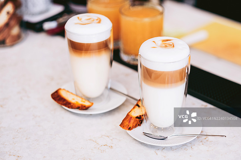 两杯玛奇朵拿铁，配有绵密的奶泡，搭配碧司奇脆饼，盛放在白色碟子上。背景是设有柔和咖啡馆灯光的白色大理石吧台。图片素材