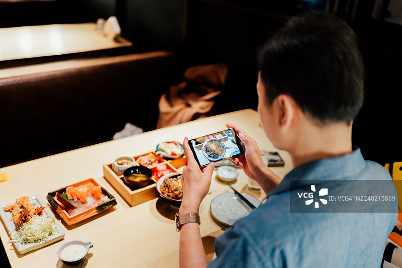 从肩膀上方拍摄的年轻亚洲商务人士在日式餐厅的餐桌旁，用智能手机拍照。她正享用午餐，并在社交媒体上分享美食照片。图片素材