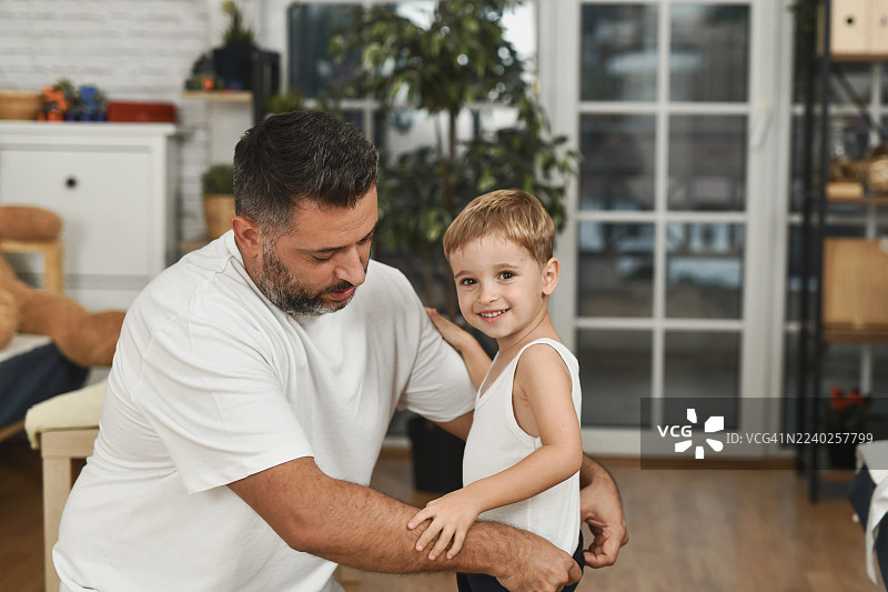 父亲在家中照顾患有水痘的孩子，在客厅为他穿衣并安慰他。图片素材