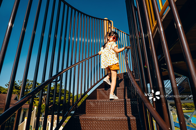 一个快乐的亚洲小女孩戴着红色太阳镜，在晴朗宜人的日子里，于户外游乐场的丛林健身器材上玩耍，享受乐趣。她微笑着看向镜头。户外乐趣。无忧无虑和自由的概念。图片素材