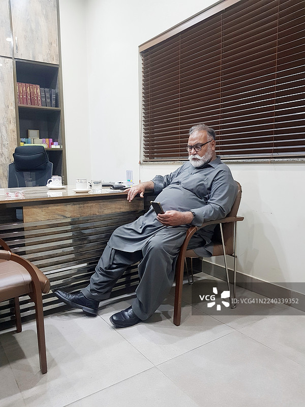 一位穿着沙尔瓦卡米兹的成熟男子坐在办公室的椅子上使用手机。图片素材