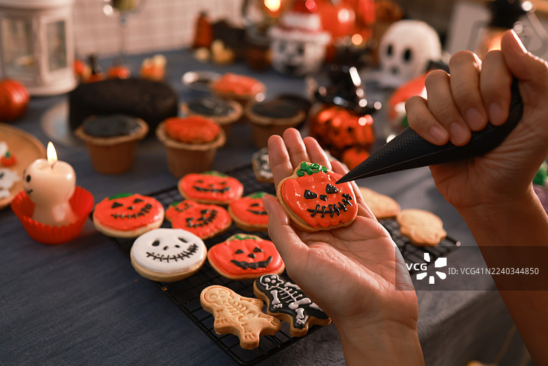 特写一位女士的手，手中拿着装饰成橙色南瓜的万圣节饼干，一位家庭主妇正在为万圣节活动准备蛋糕和饼干。图片素材