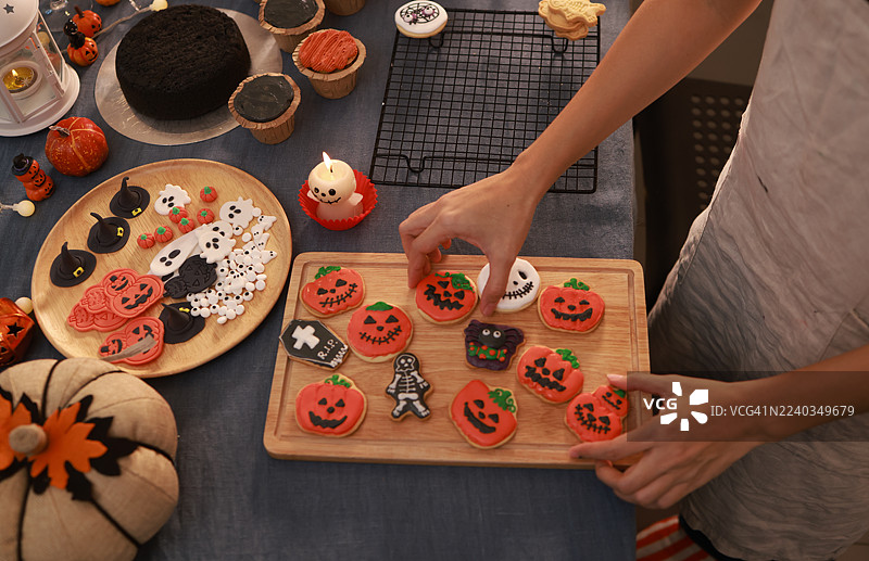 特写一位女性的手，手中拿着装饰成橙色南瓜的万圣节饼干，一位家庭主妇正在为万圣节活动准备蛋糕和饼干。图片素材