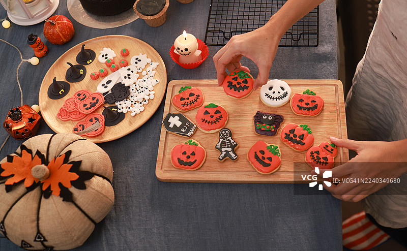 特写一位女士的手，手里拿着装饰成橙色南瓜的万圣节饼干，一位家庭主妇正在为万圣节活动准备蛋糕和饼干。图片素材