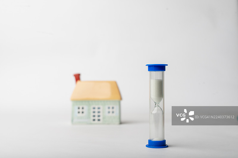 沙漏摆放在小房子模型旁边，背景简洁，突显了居家生活的时光流逝。图片素材