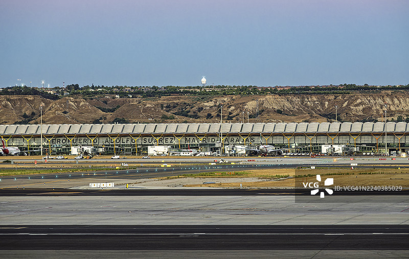 飞机在黄昏时分于马德里机场的跑道上滑行，前方是控制塔和航站楼，灯光已亮起。图片素材