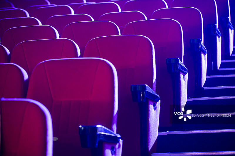 空无一人的红色天鹅绒座椅排在礼堂中，在紫色的舞台灯光下闪耀，象征着对演出或电影首映的期待。图片素材