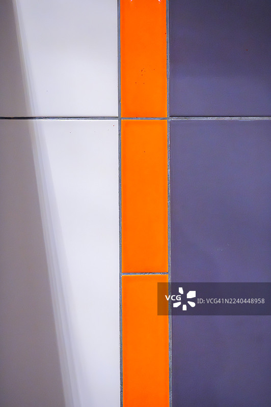 抽象几何瓷砖墙图案，展示了一条垂直的橙色条纹，分隔着白色和紫色的陶瓷表面，创造出具有光滑纹理的现代多彩背景图片素材