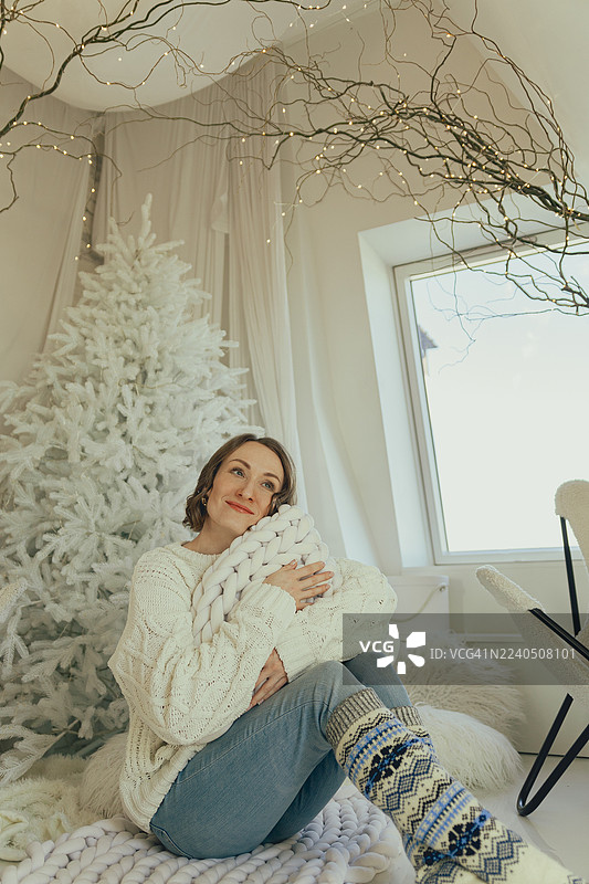一位年轻女子在节日阁楼里，抱着厚实的针织毯，依偎着白色圣诞树，营造出梦幻般的氛围。图片素材