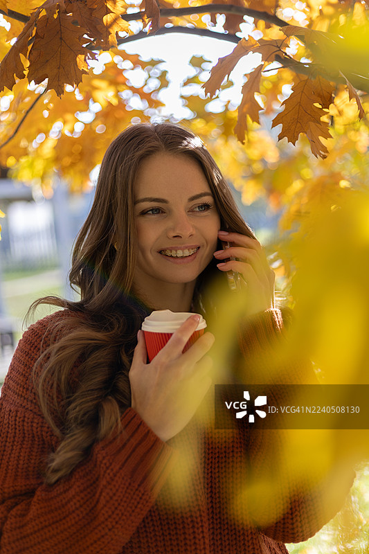 阳光明媚的日子里，一位年轻的女士微笑着在电话中交谈，手里拿着一个一次性杯子，背景是一棵黄色的橡树。图片素材