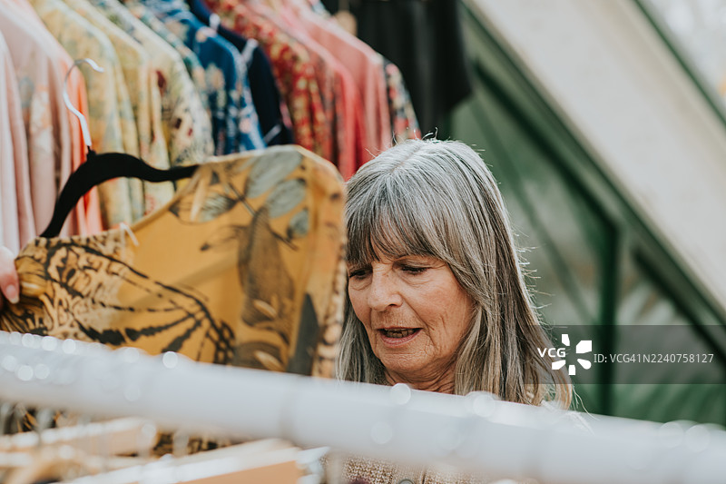 一位迷人的年长女性在一个阳光明媚的日子里，在室内市场的一个服装摊位前愉快地购物。图片素材