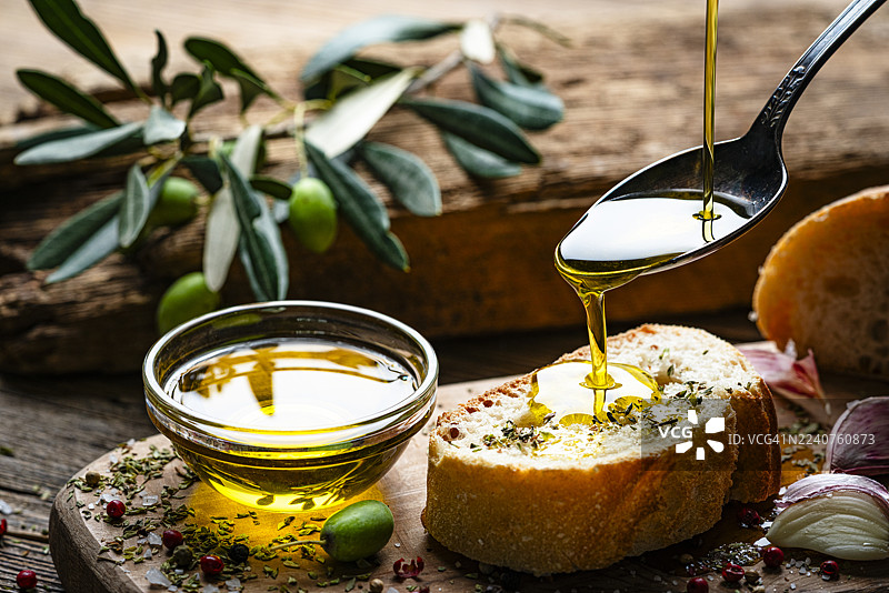 地中海美食：特级初榨橄榄油从勺子淋在乡村木桌上的面包片上。配有绿橄榄、胡椒、盐和大蒜。美味佳肴图片素材
