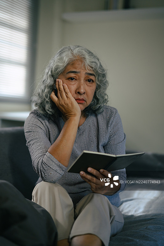 一位年长的亚洲女性坐在沙发上，手持笔记本，在柔和的自然光下陷入沉思图片素材