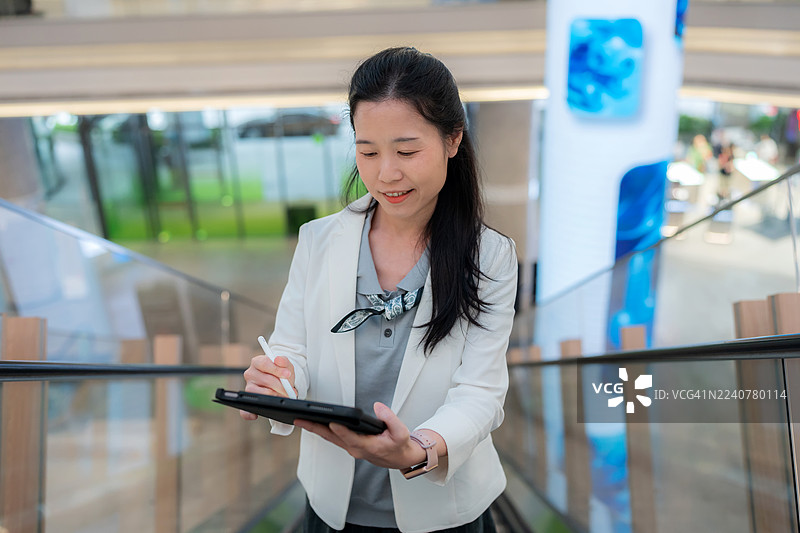 一位穿着时尚白色套装的微笑女经理站在电梯里，使用平板电脑并记录笔记。现代商务生活方式中关于激励、健康、个性化和创意自由的概念。图片素材
