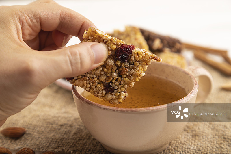 焦糖、坚果、谷物脆片格兰诺拉麦片棒，放在陶瓷盘里，背景是白木台面、米色亚麻餐巾和一杯绿茶。手部特写，侧面视角，选择性对焦。图片素材