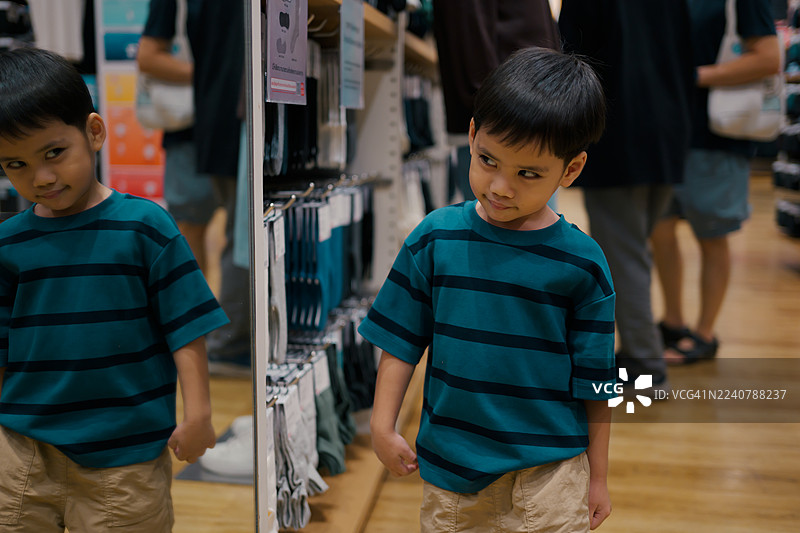 一个亚洲男孩站在服装店里，看着大镜子里自己的倒影，购物时显得好奇。图片素材
