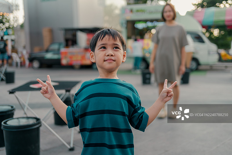年轻男孩在街头户外嘉年华上对着镜头微笑，双手比出象征和平的手势，旁边停着餐车。图片素材