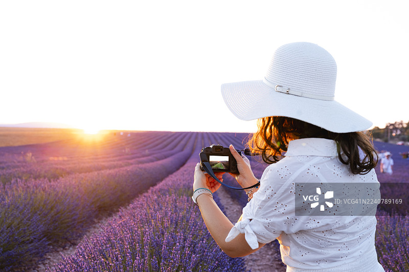 法国瓦朗索勒，一位身着白裙、头戴宽檐帽的女士在日落时分拍摄薰衣草田。图片素材