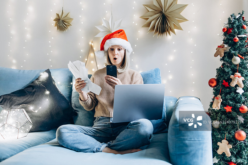 身着圣诞帽的震惊女子坐在舒适的沙发上，旁边放着笔记本电脑和收据，在圣诞季看着她的智能手机。概念为假期购物、在线消费、预算、圣诞开销。图片素材