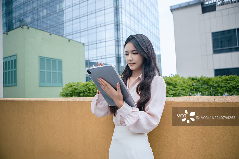 一位专注的亚洲女商人站在现代摩天大楼露台上使用iPad。职业女性企业家在户外使用数字设备，背景是城市天际线，代表着生产力、专注和现代商业生活方式。图片素材