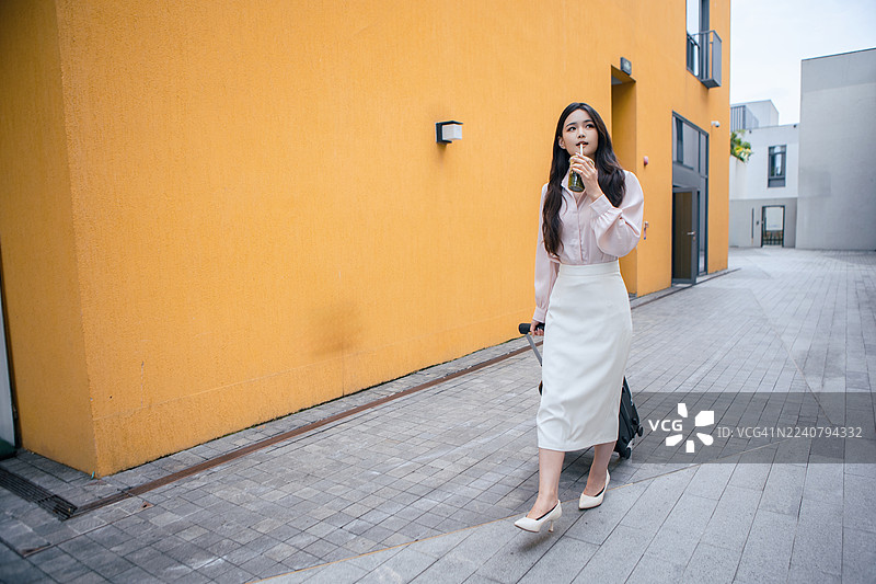 亚洲女商人正在城市旅行，沿着黄墙街道行走，同时在寻找酒店。一位专业的女性创业者，提着行李，探索城市环境，图片素材