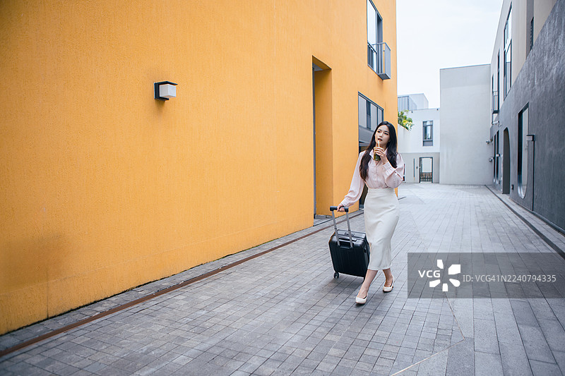 亚洲女商人正在城市旅行，走在一条黄墙街道上寻找酒店。一位专业的女企业家，带着行李，探索城市环境，图片素材